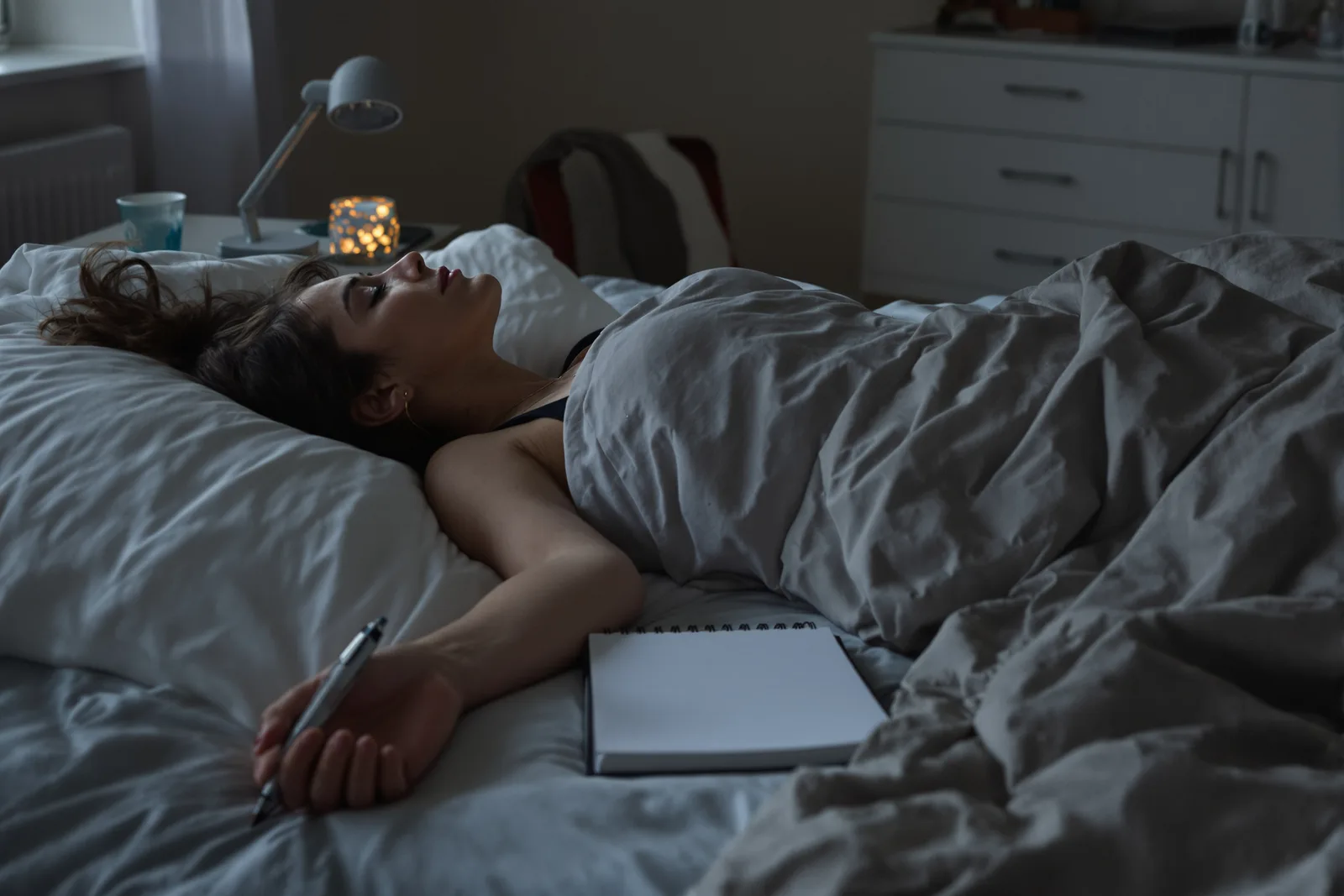 Woman lying in bed in the evening with eyes closed, pen and notebook beside her, a quiet moment for a posthypnotic suggestion