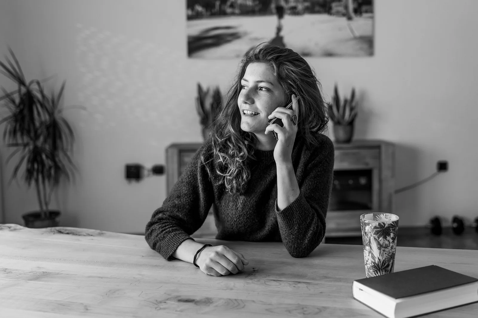 Woman smiling on the phone at a wooden table next to a glass and a book: connection instead of comparison