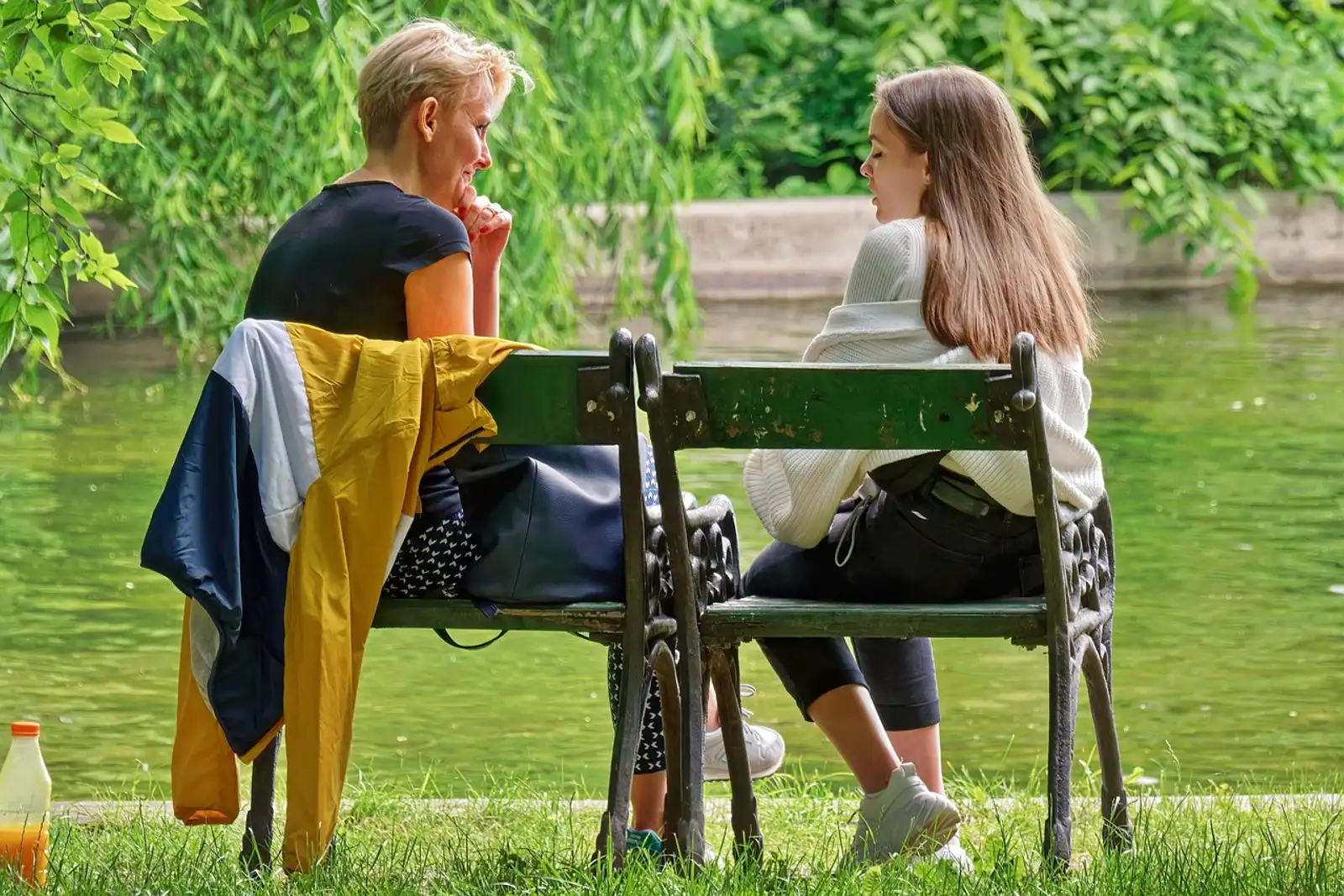 Two women talking on park benches by a pond, because talking openly helps you stop feeling like a fraud at work