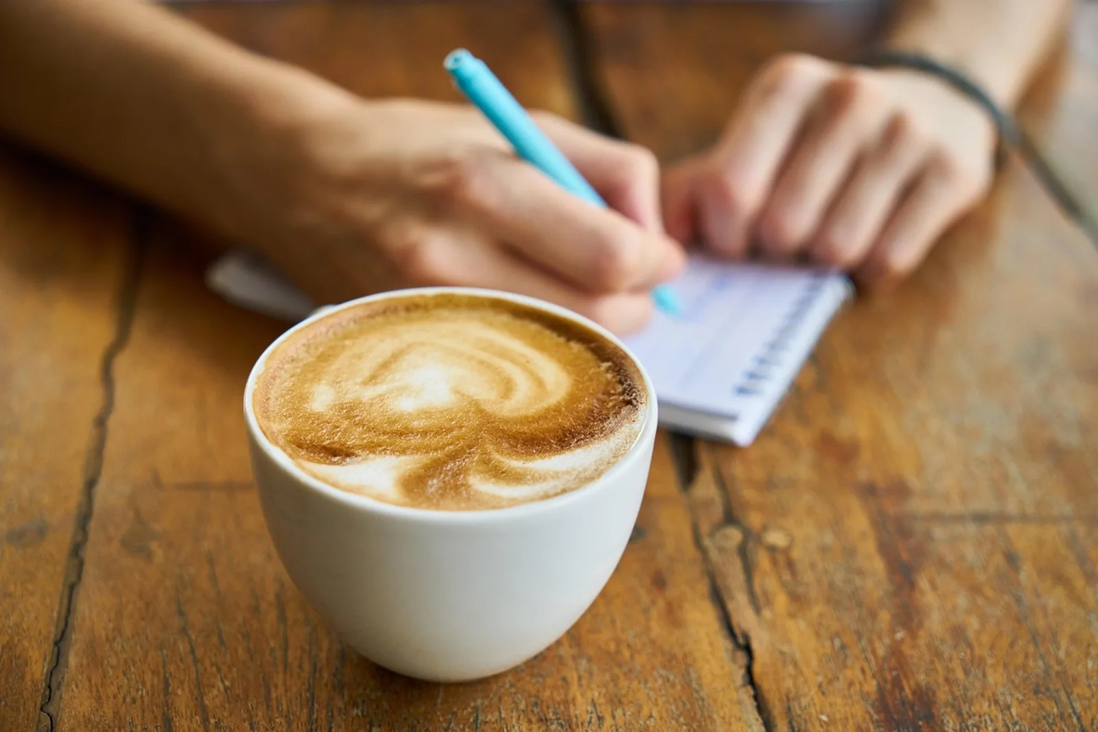 Hands writing a sentence with a light blue pen next to a cappuccino: a small ritual to quiet the inner critic