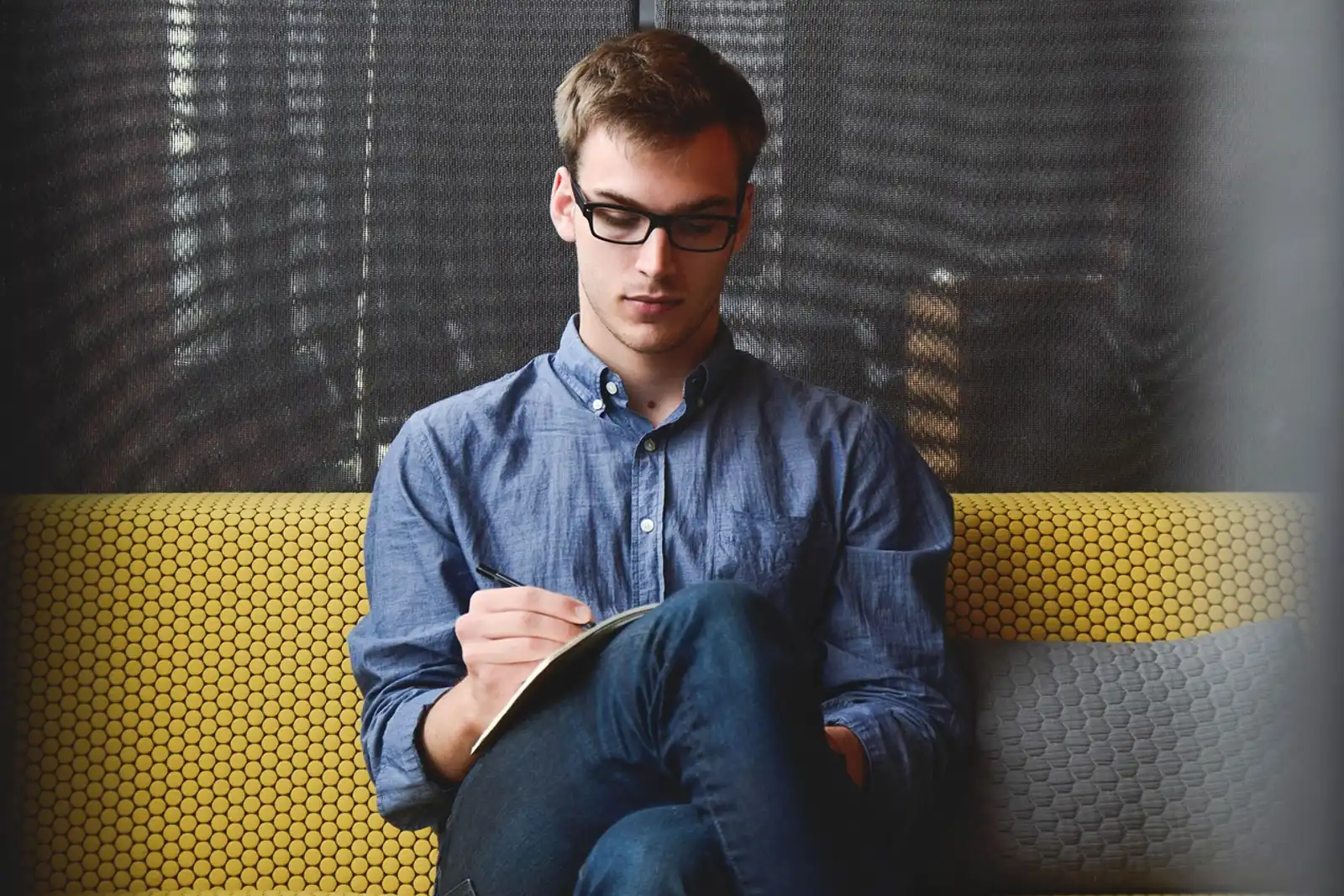 Man with glasses sitting on a sofa writing in a notebook: writing down self-doubt to overcome it